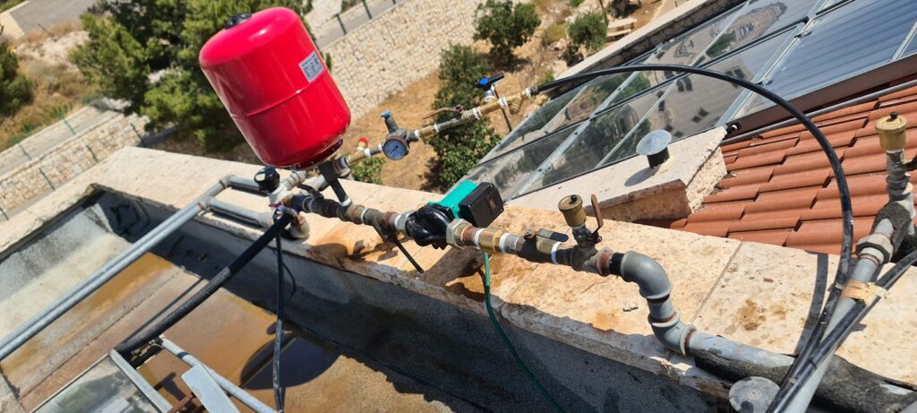 משאבת סחרור מים חמים עם מיכל התפשטות אדום על גג בניין משותף ליד קולטי שמש